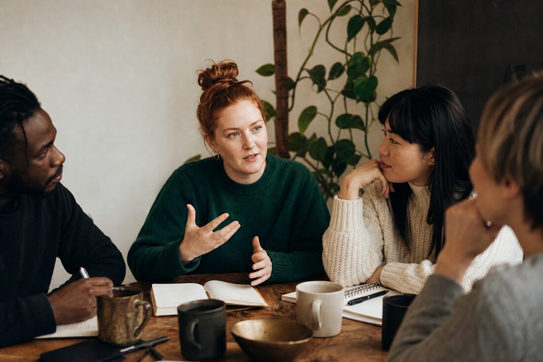 A small group in conversation around a wooden table with notebooks and mugs